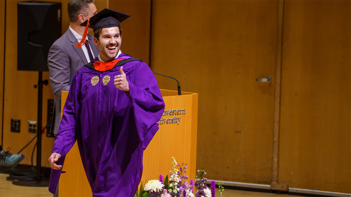A graduate gives a thumbs-up gesture as he walks across the stage.